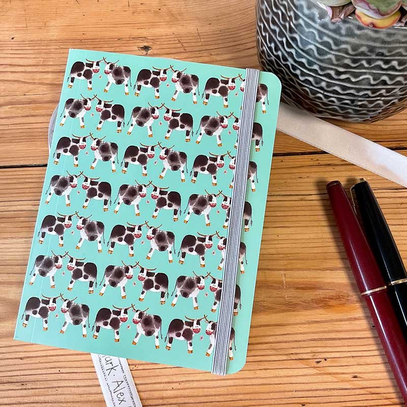 Alex Clark Dairy Cows Small Chunky Notebook front cover on a wooden table with pens